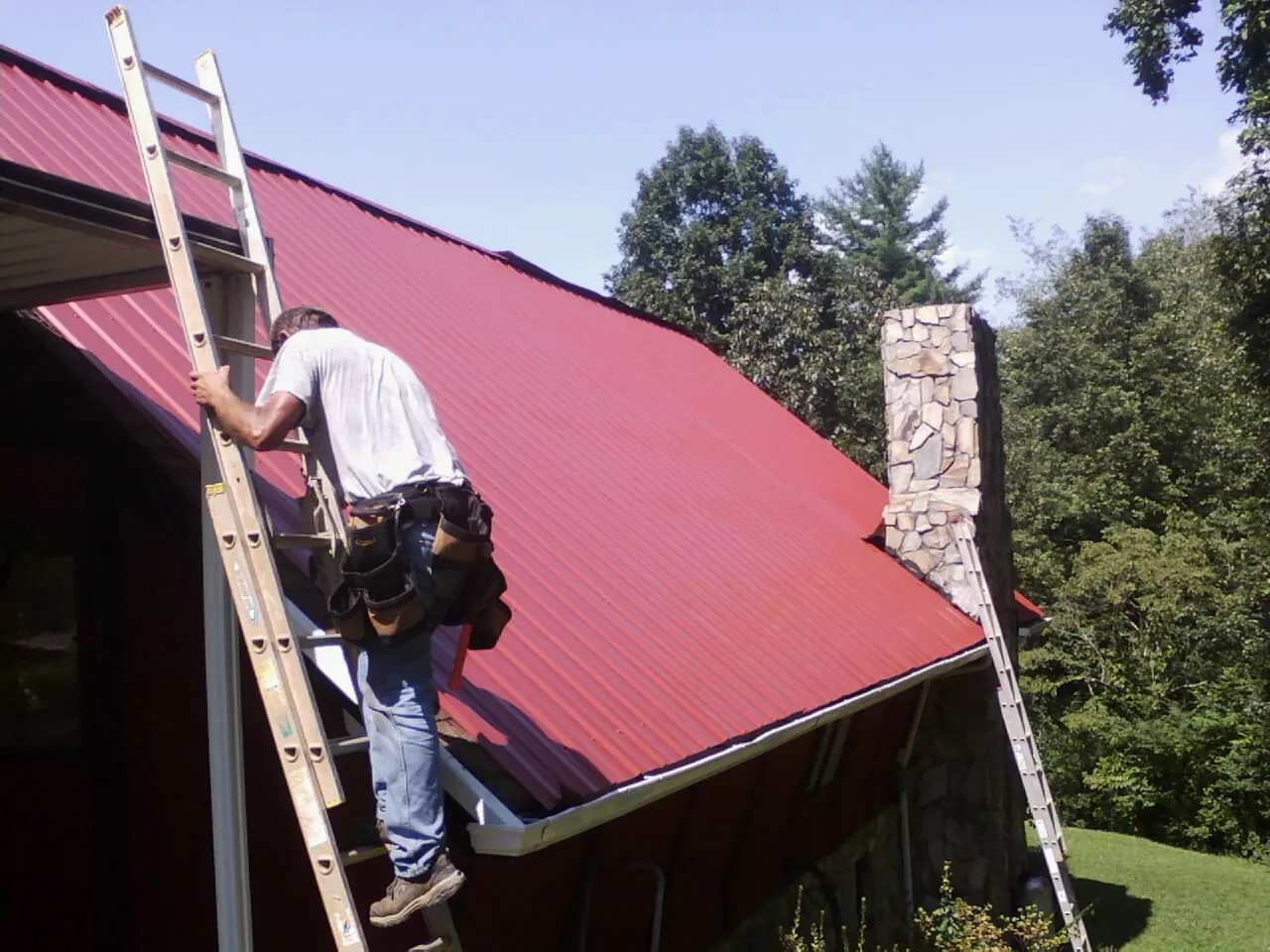 Roofer on a ladder beside a red metal roof and stone chimney in Garden Valley